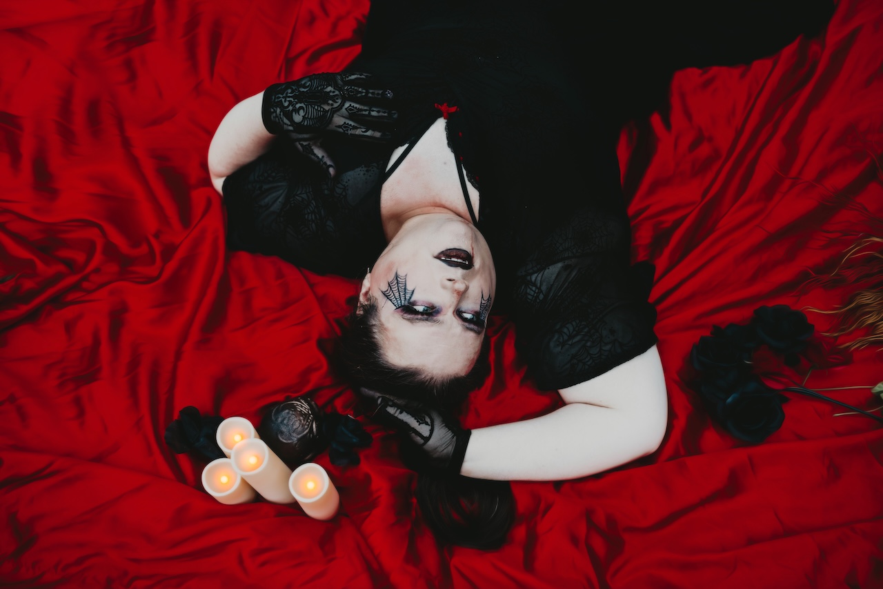 Jenni laying down on a luxurious red sheet. She has spiderweb eyeliner makeup, sheer mesh gloves, and a wrap around dress. All black everything, even her lip color.